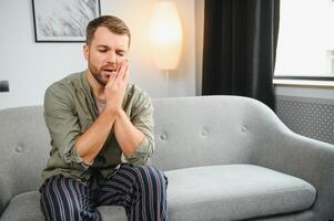 Problems with teeth. Toothache. Upset caucasian guy is sitting on the couch at home, holding his hand to his cheek, having a toothache, needs a dentist consultation, sad facial expression. photo