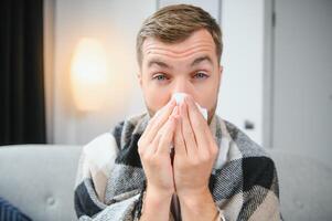 Young man suffering from a common cold and flu or allergy sit at home wrapped in blanket and wipe his nose with tissues while he have a strong headache pain, healthcare concept. photo
