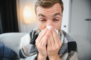 Young man suffering from a common cold and flu or allergy sit at home wrapped in blanket and wipe his nose with tissues while he have a strong headache pain, healthcare concept. photo