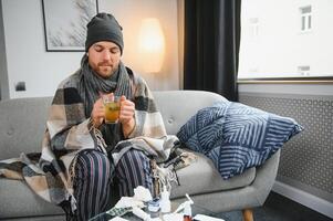 Sick bearded man who has bad cold or seasonal flu sitting on couch at home. Guy with fever wearing warm plaid shivering with worried face expression. photo