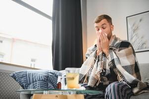 Young man suffering from a common cold and flu or allergy sit at home wrapped in blanket and wipe his nose with tissues while he have a strong headache pain, healthcare concept. photo