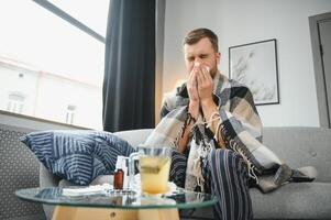 Young man suffering from a common cold and flu or allergy sit at home wrapped in blanket and wipe his nose with tissues while he have a strong headache pain, healthcare concept. photo