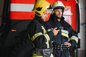 Two professional firefighters with uniforms and protective helmets posing infront of a firetruck. photo