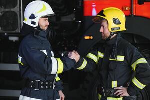 Two firefighters in protective clothing in helmets with fire engine, friendly handshake photo