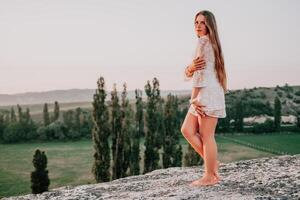 Happy woman in white boho dress on sunset in mountains. Romantic woman with long hair standing with her back on the sunset in nature in summer with open hands. Silhouette. Nature. Sunset. photo