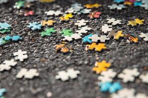 A background image of pile of mixed colorful jigsaw Puzzle pieces set scattered on the gray asphalt pavement on the street. Messy chaotic concept. Close up. Selective focus photo
