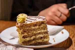 Slice of tasty homemade delicious sweet Esterhazy cake with nuts and cream. Piece of Esterhazy Torte served on the white plate with napkin. Young womans hand on the blurred background Selective focus photo