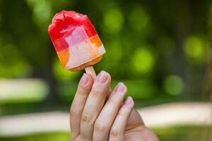 Close-up of a vibrant colorful red yellow orange bitten ice cream with popsicle stick held in a hand of unrecognizable child. Sunny summer day in park. Blurred background. Copy space. Selective focus photo