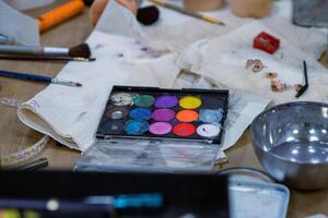 Messy colorful creative table with makeup palette, paints, brushes, pencil sharpener, paintbrushes set and bowl of water, paper tissues on wooden table. Stage make up. Creative chaos. Selective focus photo
