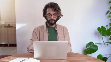 A man with glasses and a beard is sitting at a table at home using a laptop computer video