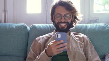 Happy man with a beard is sitting on a couch using a cell phone on a call video