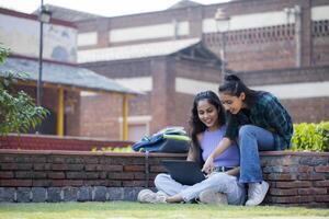 Two College Classmates Are On the University Premises, One of Whom is Pointing Her Finger At the Laptop of a Sitting Girl. photo