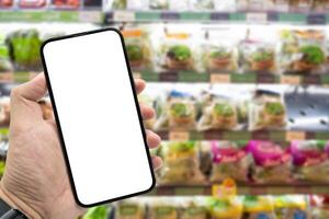 Hand holding blank screen smartphone for copy space on blurred Miscellaneous Product shelf. e-commerce app shopping mockup. social media, payment and shopping online concept. photo