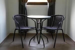 The faux rattan table and chair set consists of 2 chairs and a black round table placed next to the window with black curtains in the bedroom for sitting or working. photo