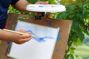 An artist is using a paintbrush to paint poster paint in green paint to paint on a painting while out in the field. soft and selective focus. photo