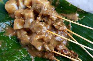 Delicious sate padang dish from Indonesia on top of paper plate and green banana leaf background. Beef skewers with sauces and ketupat rice cakes. photo