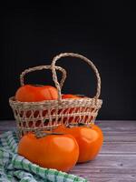 Fresh ripe persimmons in a basket and on a wooden table with a black wall background. Space for text. Close-up photo. Concept of health fruit photo