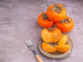 Fresh ripe persimmons with slices of persimmon on a plate with cutlery and whole fruit on gray stone background. Space for text. Close-up photo. Concept of health fruit photo