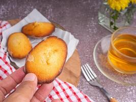 Hand holding a piece of madeleine on lemon glaze madeleines on a plate with a cup of tea placed on a gray stone background. Madeleine - homemade traditional French small cookie photo