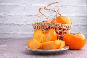 Fresh ripe persimmons with slices of persimmon on a plate and whole fruit in a basket with a white brick wall. Space for text. Close-up photo. Concept of health fruit photo