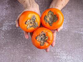 Three persimmons on the palm senior woman with a gray stone background. Close-up photo with copy space for text. Top view. Concept of aged people and health fruit