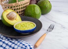 Organic avocado with seed, avocado halves, and fresh guacamole in a bowl on black plate with cloth and cutlery on marble background. Top view. Space for text. Flat lay style.Concept of healthy fruit photo