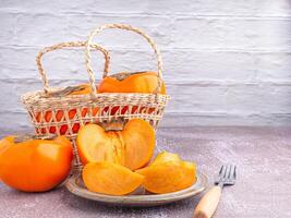 Fresh ripe persimmons with slices of persimmon on a plate with cutlery and whole fruit in a basket with a white brick wall. Space for text. Close-up photo. Concept of health fruit photo