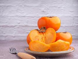 Fresh ripe persimmons with slices of persimmon on a plate with cutlery and whole fruit in a basket with a white brick wall. Space for text. Close-up photo. Concept of health fruit photo