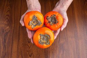 Three persimmons on the palm senior woman with a wooden table background. Close-up photo with copy space for text. Top view. Concept of aged people and health fruit