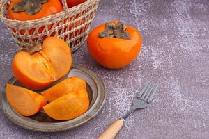 Fresh ripe persimmons with slices of persimmon on a plate with cutlery and whole fruit in a basket with a white brick wall. Space for text. Close-up photo. Concept of health fruit photo