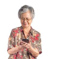 Portrait of an elderly Asian woman holding and looking at a smartphone and smiling while standing with a white background. Space for text. Concept of aged people and technology photo