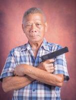 Portrait of an elderly Asian man holding a short gun and looking at the camera while standing with a brown background. Space for text. Concept of aged people and sport photo