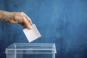 Closeup of a hand casting a white paper ballot into a transparent box on a blue background photo