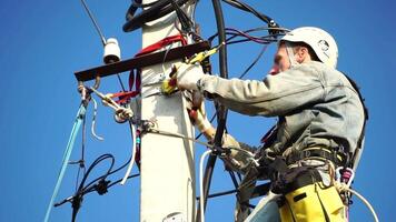 Skilled electrician in helmet fixes wires standing on ladder near high pole against blue sky on summer day backside view. Electrical service and mounting on the pole. Slow motion video