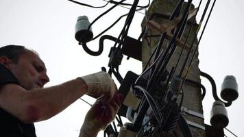 Skilled electrician fixes wires standing on ladder near high pole against blue sky on summer day backside view. Electrical service and mounting on the pole video