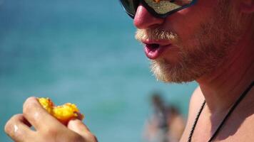 Man eating peach. Bearded man in sunglases eating peach on the beach. Happy man is smiling and resting at sea. Bearded guy enjoying fruits on summer vacation. Vegetarian and healthy lifestyle concept. video