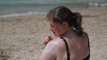 Fair skinned woman applies sunscreen to shoulder on sea beach on sunny day. Protection from harmful effects of ultraviolet rays while relaxing on ocean coast with light waves. video