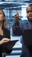 Vertical System administrators in data center overseeing server operations using notebook and tablet. Colleagues brainstorming in server hub, ensuring optimal performance and data integrity, camera A video