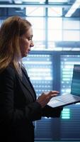 Vertical Woman walking in high tech facility, using laptop to patch network issues leading to slow data transfer rates. Employee in data center fixing errors affecting servers performance, camera B video