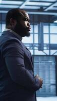 Vertical African american technician walking in data center, monitoring systems to identify errors. Man in server hub location doing diagnosis, looking for infrastructure bugs in need of repairing, camera A video