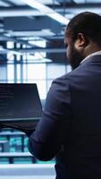 Vertical Computer scientist in server farm using laptop to patch network issues leading to slow data transfer rates. IT expert in data center establishment fixing errors affecting supercomputers, camera A video
