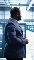 Vertical African american technician walking in data center, monitoring systems to identify errors. Man in server hub location doing diagnosis, looking for infrastructure bugs in need of repairing, camera B video