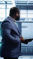Vertical Admin doing yearly checkup using notebook to prolong data center electronics life span and avoid downtime. Man walks in server room, monitoring server infrastructure components using laptop, camera B video