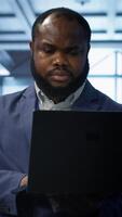 Vertical Computer scientist in data center using diagnostic tools and system monitoring software on laptop to identify cause of errors. IT specialist in server farm establishment repairing equipment, camera A video