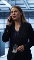 Vertical Software engineer talking on smartphone in data center, coordinating server maintenance tasks. Woman surrounded by server racks and high tech equipment, speaking during telephone call, camera A video