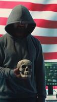 Vertical American man in a dark hood making death threats with human skull concept, emphasizing violence and malicious intent. Governmental hacker inflicting terror and panic for rival ideologies. Camera A. video