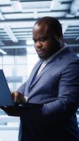 Vertical African american IT operator doing maintenance using laptop to prolong data center equipment life span. IT professional in server hub workspace monitoring server mainframes using notebook, camera B video