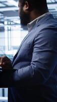 Vertical Software developer in server hub checking recovery plan on laptop, monitoring hardware energy consumption. African american worker uses notebook in data center, ensuring rigs are functioning, camera B video