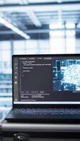 Vertical Focus on laptop in data center with artificial intelligence brain used by engineers in blurry background. Close up of device displaying AI deep learning software in server farm, camera B video