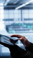 Vertical System administrator monitoring server room, doing checkup, inputting data on tablet of needed fixes. IT support employee working in data center facility housing storage rackmounts, camera B close up video